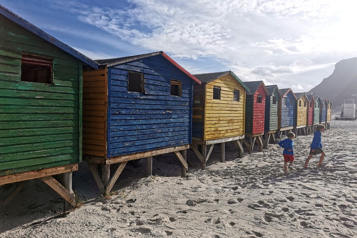 Two boys walking next to the colourful beach huts on Muizenberg Beach in Cape Town, one of the best beaches in Cape Town.