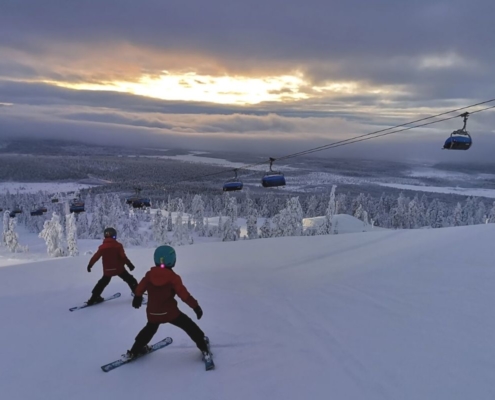 Two kids skiing down a ski slope in Levi Ski Resort in Finland with sun poking through the clouds.