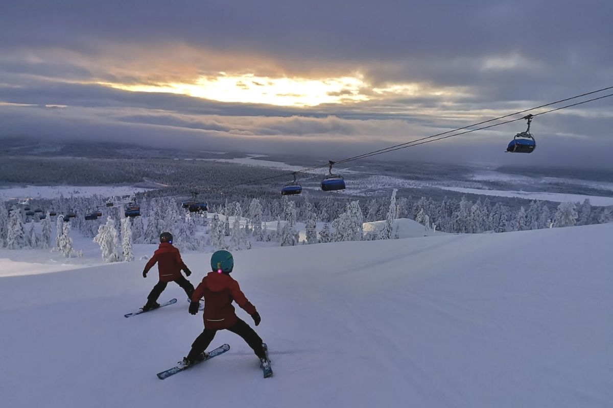 Two kids skiing down a ski slope in Levi Ski Resort in Finland with sun poking through the clouds.