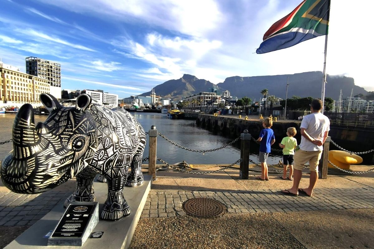 Father and two sons standing at the V&A Waterfront in Cape Town looking towards Table Mountain with a black and white rhino in foreground.