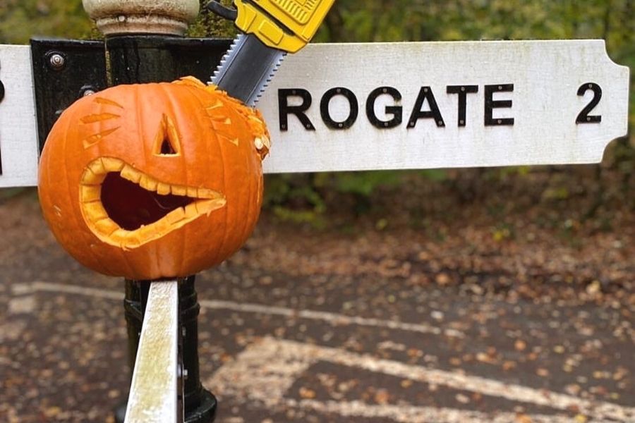 Halloween decoration of a pumpkin with a chainsaw through it around a road sign with Rogate on it.