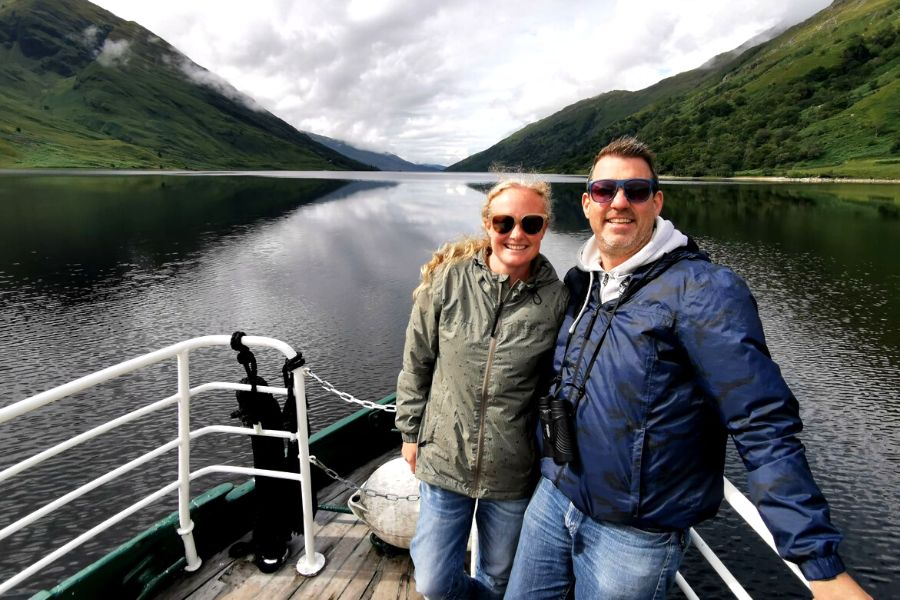 Couple wearing sunglasses and TOG24 craven packaway jackets on a boat on a lake in Scotland with mountains in background.