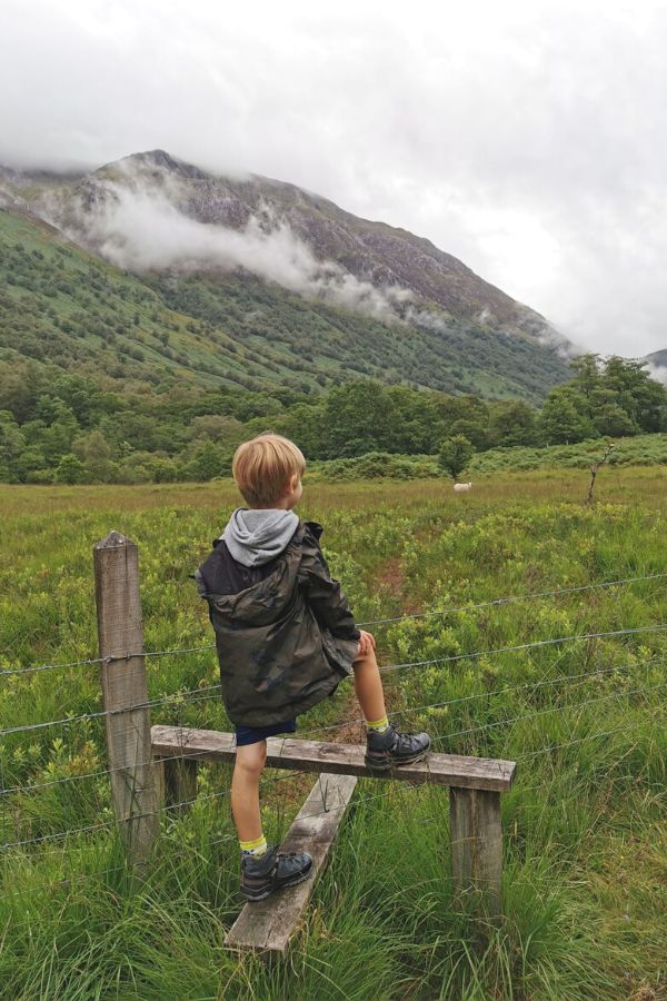Young boy in a TOG24 waterproof jacket standing on a stile looking at Glen Nevis with a line of cloud cover.
