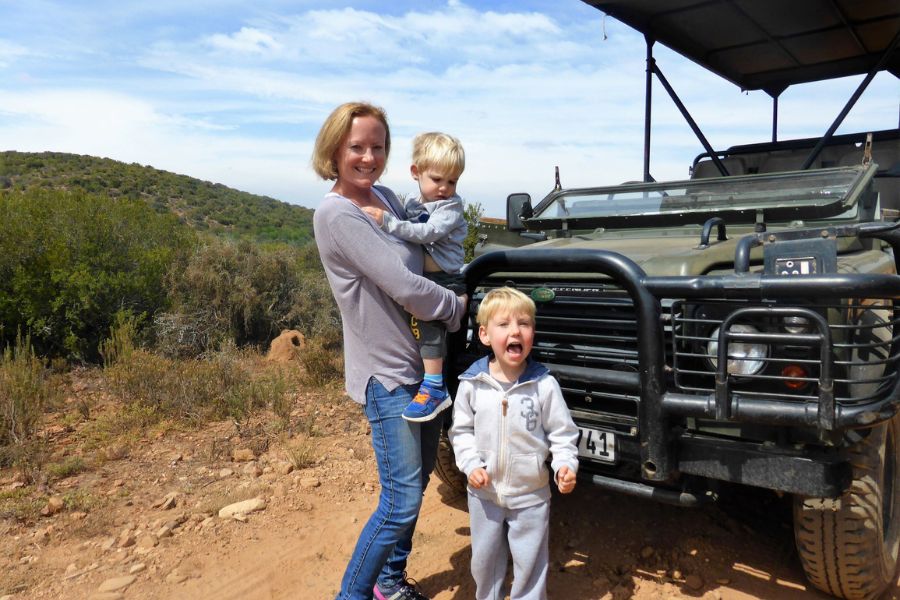 Mum carrying a toddler with another small boy in front of a safari jeep on a malaria free safari in South Africa.