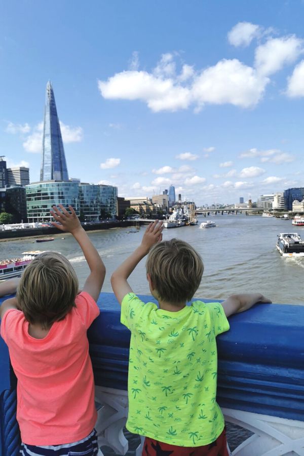 Two boys standing on Tower Bridge waving to boats as they pass under the bridge.