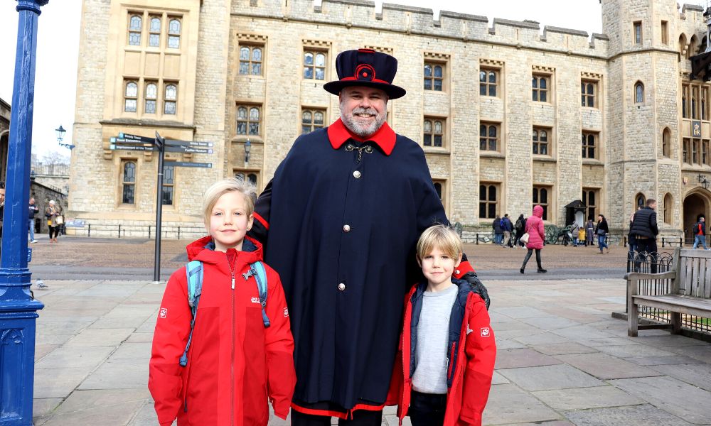 Two young boys in red coats standing either side of a Yeoman at the Tower of London with kids.