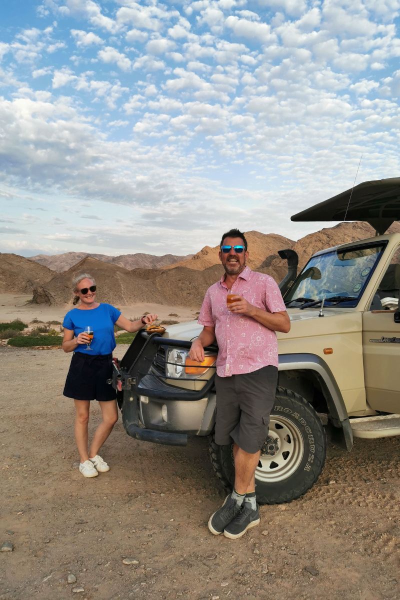 Couple enjoying sundowner drinks by a jeep in the Hoanib Valley in Namibia on an epic two week Namibia road trip.