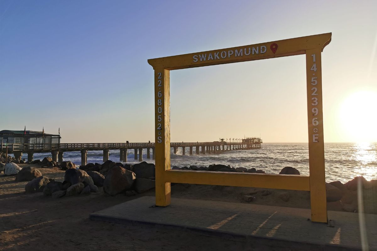 Large yellow frame sign of Swakopmund with Swakopmund pier in the background at sunset.
